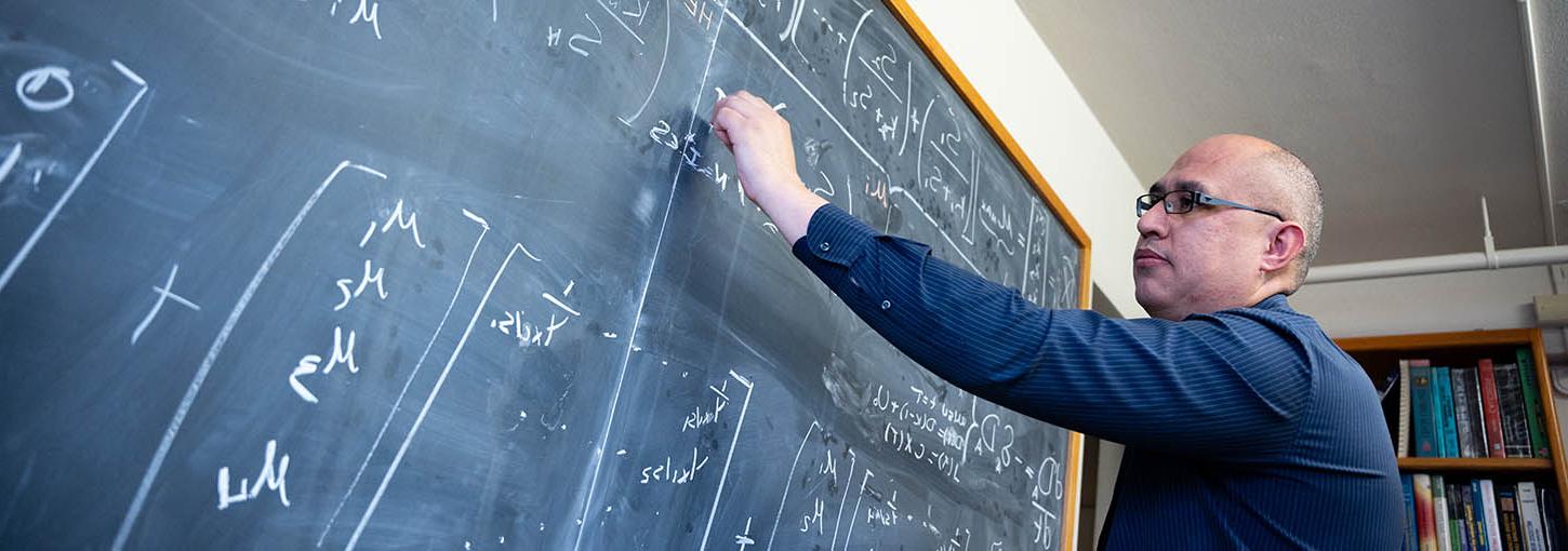 一个戴眼镜穿蓝衬衫的人在黑板上写数学公式.